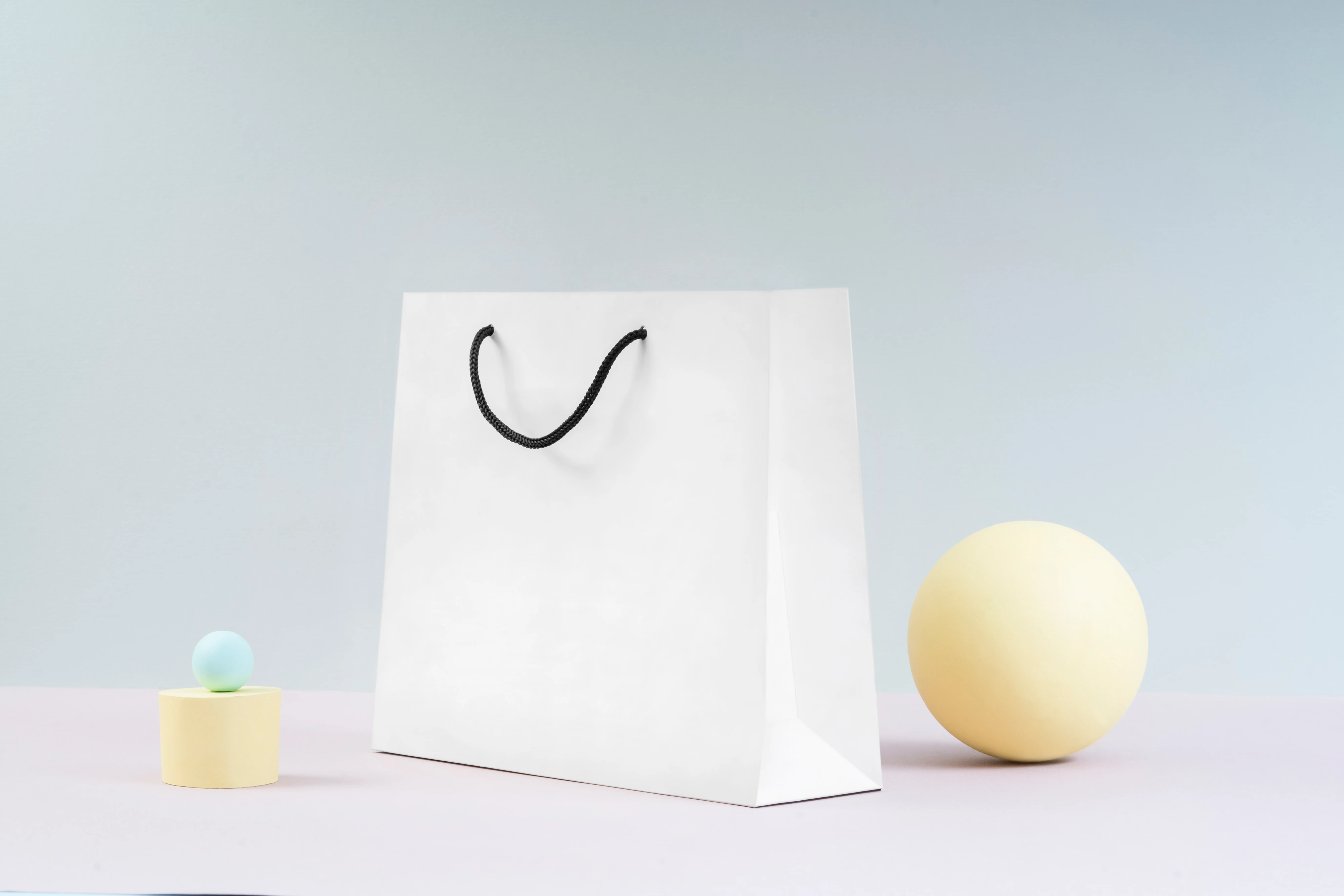 Brand still-life: a white shopping bag on a pale stage with soft pastel props, the quiet backdrop chosen for product to read against.