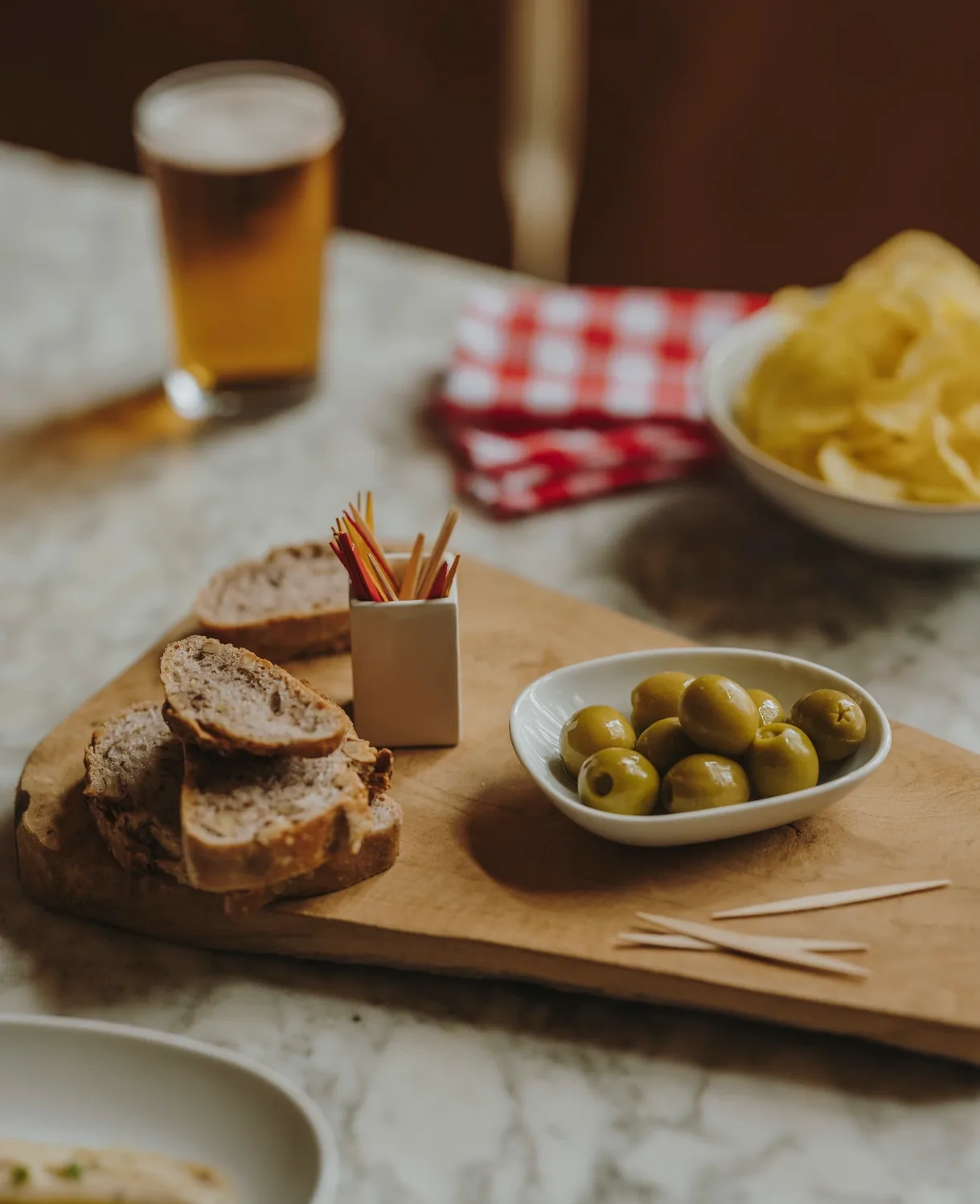 Wooden board with bread, olives in a bowl, toothpicks, beer and crisps on a marble table