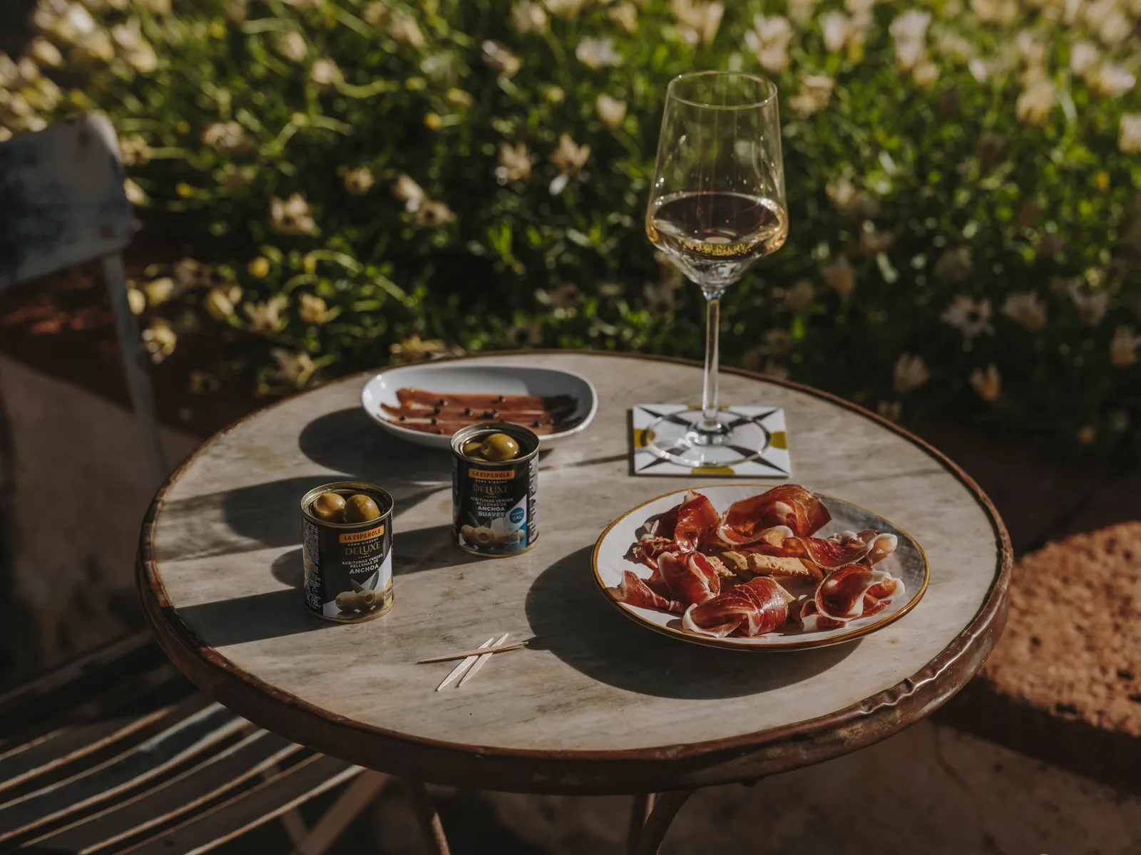 Sunlit terrace table with anchovies, prosciutto, wine and La Española tins, garden in background